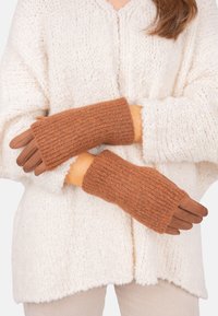 Person, der braune Strickhandschuhe mit Wildlederfingern und eine cremefarbene strukturierte Strickjacke trägt, die die Arme auf Hüfthöhe überkreuzt.
