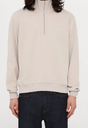 Person trägt beige Viertel-Reißverschluss-Sweatshirt mit "Tiger of Sweden" Logo und dunkelblaue Jeans, steht vor weißem Hintergrund.
