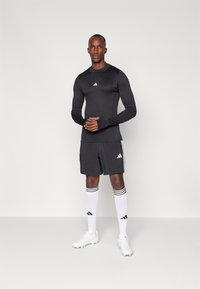 Schwarzes langärmliges Sportoberteil mit weißem Logo, kombiniert mit schwarzen Shorts, weißen knielangen Socken mit schwarzen Streifen und weißen Sportschuhen.