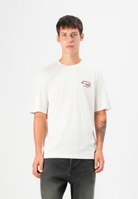 Kortærmet t-shirt i bomuld i off-white med et rødt grafisk design på venstre bryst. Har en rund halsudskæring og en afslappet pasform.