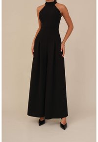 Vrouw in een mouwloze zwarte halter jumpsuit met wijde broekspijpen en zwarte puntige hakken tegen een neutrale achtergrond.
