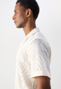 Witte gehaakte poloshirt met golfpatronen, korte mouwen en een kraag. Het materiaal lijkt lichtgewicht en textuur te hebben.