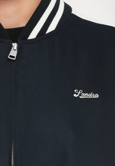 Veste zippée bleu marine avec col côtelé à rayures blanches, arborant le logo brodé "Sandro" sur le côté gauche. Texture du tissu lisse.