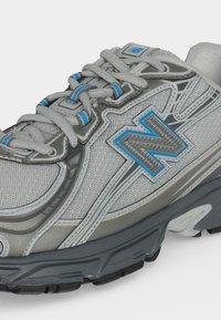 Γκρι και μπλε αθλητικό παπούτσι New Balance με πάνω μέρος από δίχτυ, λεπτομερή ραφές, χοντρή σόλα και δεμένα κορδόνια σε λευκό φόντο.