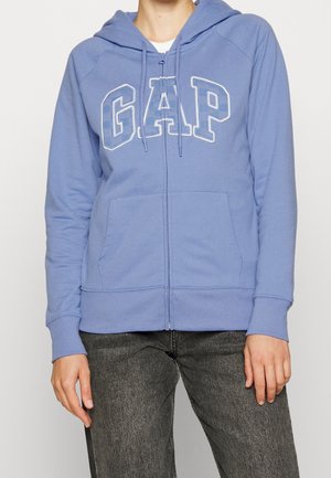 Sweat à capuche zippé bleu avec un logo "GAP" blanc. Dispose d'une capuche, de poignets côtelés et de poches avant. Fabriqué en tissu doux avec une texture lisse.