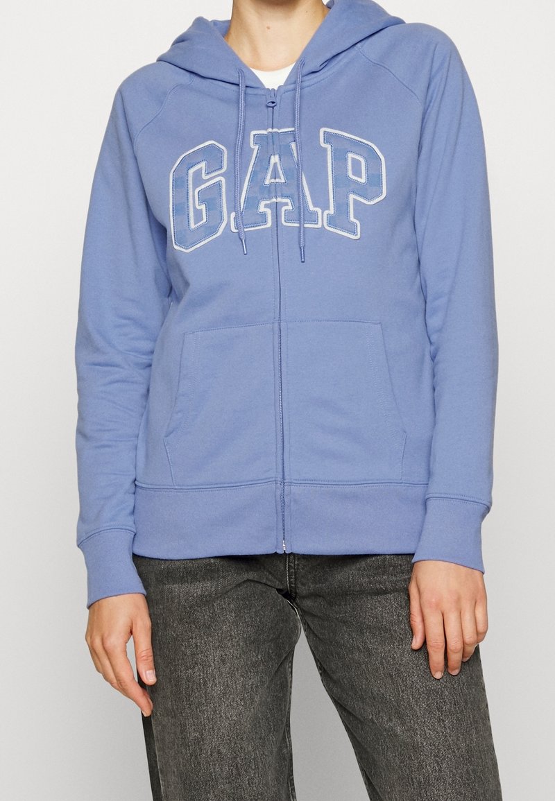 Sweat à capuche zippé bleu avec un logo "GAP" blanc. Dispose d'une capuche, de poignets côtelés et de poches avant. Fabriqué en tissu doux avec une texture lisse.
