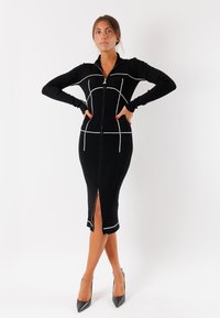 Abito nero a coste con pattern a griglia bianca, chiusura con zip e spacco frontale. Silhouette aderente con maniche lunghe e colletto a finta. Indossati tacchi alti.