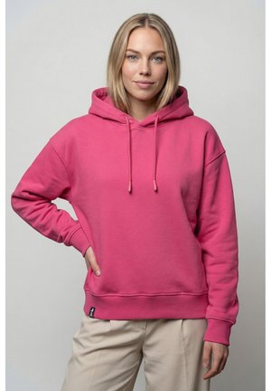 Junge Frau mit blonden Haaren, die einen leuchtend pinken Hoodie und beige Hose trägt und mit einer Hand in der Hüfte vor einem einfarbigen Hintergrund steht.