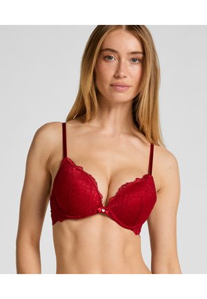 Soutien-gorge en dentelle rouge avec armatures, présentant une bordure festonnée, des bretelles rouges réglables et un petit nœud décoratif au centre.