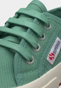Close-up van een groene canvas sneaker met bijpassende veters, zilveren veterogen, witte rubberen zool en een Superga-logotag aan de zijkant.