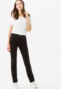 Pantaloni neri slim-fit in cotone, con chiusura frontale a bottone, cinque tasche e una texture liscia. Abbinati a una maglietta bianca larga e sneakers bianche.