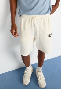 Shorts de sport de couleur crème avec poches latérales et taille élastique, portant un logo "Converse" noir. Assortis avec des baskets beiges.
