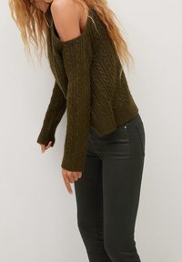Mujer vestida con un suéter de punto trenzado en color verde oliva y pantalones de cuero negros, de pie contra un fondo beige.