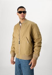 Tanan bomber jakna s sprednjim zadrgo, dvema nagnjenima žepoma in rebrastimi manšeti. Model nosi črna pravokotna sončna očala in modre kavbojke.