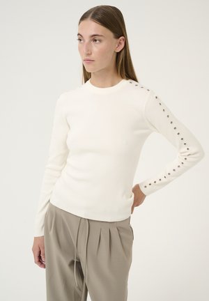 White knit sweater with a round neckline and long sleeves, featuring decorative metal buttons along the sleeve seams. Paired with light beige trousers.
