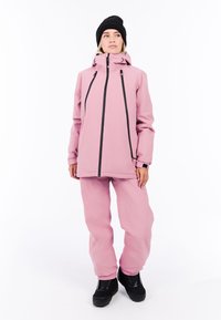 Roze waterdichte jas met zwarte ritsen en verstelbare koorden, gecombineerd met bijpassende roze broek en zwarte schoenen. Zachte textuur, relaxed fit.