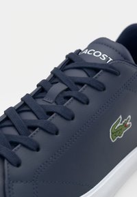 Baskets Lacoste bleu marine avec lacets foncés, logo crocodile vert sur le côté et lettrage blanc "LACOSTE" sur la languette, semelle blanche visible.