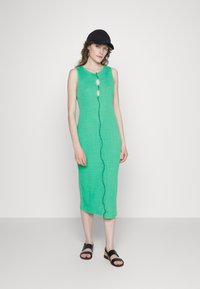 Vestido de linho verde com acabamento texturizado e botões frontais com aberturas em forma de olho-chave. Combinado com sandálias pretas e um boné.