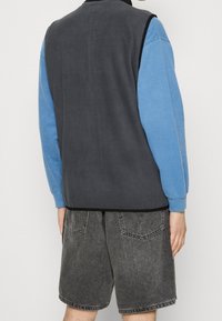 Fleeceväst i mörkgrått över en ljusblå långärmad skjorta. Har rund hals, lös passform och är kombinerad med grå denimshorts.