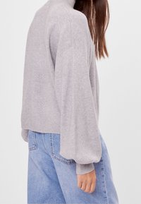 Grijze, zachte gebreide trui met een hoge kraag en ballonmouwen, gecombineerd met lichtblauwe spijkerbroek. Het ontwerp is ontspannen en oversized.