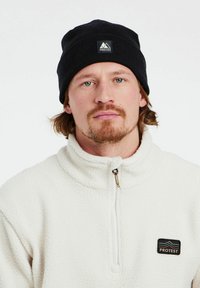 Sort strikket hue med et bjerglogo og en hvid fleece zip-jakke. Jakken har en høj krave og et lille brandmærke.