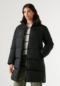 Melna pūšamā jaka ar augstu apkakli, pogu aizdari, quilted dizainu un sānu kabatām. Valkāta virs gaiši krāsotas svītrainas kreiķa.