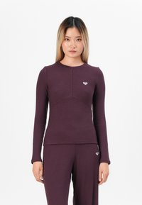 Langermet topp i ribbet, dyp lilla stoff med rund halslinning. Har et hjerteemblem på brystet og en tettsittende silhuett.
