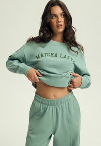Jasnozielony krótki sweter z napisem "MATCHA LATTE", zestawiony z dopasowanymi zielonymi dresami; oba wykonane z miękkiego materiału i mają elastyczny pas.