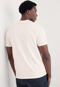 Camiseta de algodón de manga corta, color beige claro, con cuello redondo, textura suave y corte relajado, vista desde atrás contra una pared neutra.