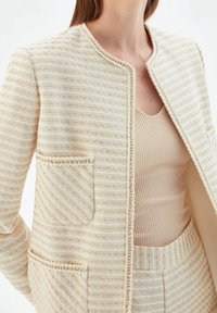 Z teksturowanym kremowym żakietem z złotą nitką, okrągłym dekoltem i bez guzików. Posiada dwie przednie kieszenie z kontrastowym wykończeniem. Nosi się go na beżowej bluzce.