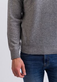 Maglione grigio lavorato a maglia con polsini e orlo a coste, indossato sopra jeans di denim blu. Primo piano mostra la trama del materiale e una mano che tiene i jeans.