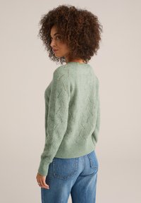 Maglione lavorato a maglia verde chiaro con un motivo a onda, maniche lunghe e polsini a costine. Indossato con jeans blu a vita alta. Tessuto testurizzato.