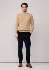 Muškarac stoji naslonjen na jednoličnu pozadinu, obučen u bež pulover, bijelu košulju s ovratnikom, crne hlače i smeđe kožne cipele, s jednom rukom u džepu.