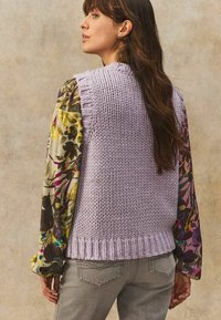 Gilet en lilas tricoté avec des bords côtelés texturés porté sur un haut à manches fleuries présentant un mélange de couleurs. Un jean gris complète la tenue.