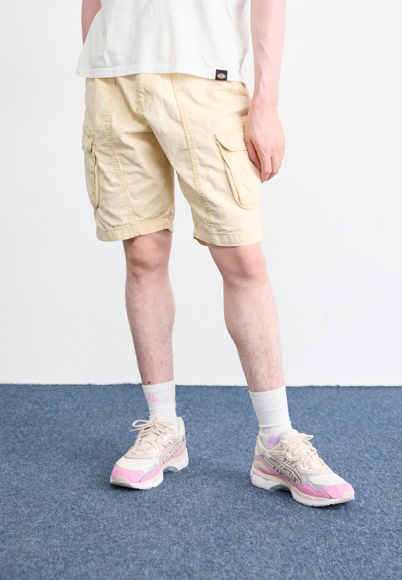 Pantaloni scurți cargo de culoare bej deschis, cu două buzunare laterale, asortați cu adidași roz deschis și crem și șosete albe până la glezne.