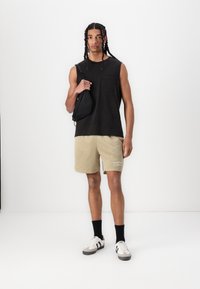 Sort ærmeløs skjorte, beige mesh shorts, sorte sokker, hvide og grå sneakers. Modellen holder en lille sort taske. Enkel, afslappet outfit.