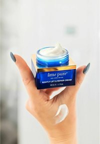 Blå glasburk med guldock, märkt "âme pure Nightly Lift & Repair Cream," fylld med vit kräm. Kräm på hand i förgrunden.