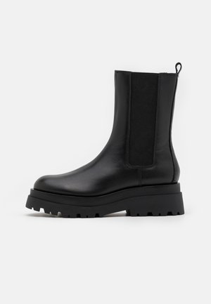 Botte à cheval en cuir noir avec semelle épaisse à crans, panneau latéral élastique et tirant sur fond blanc.