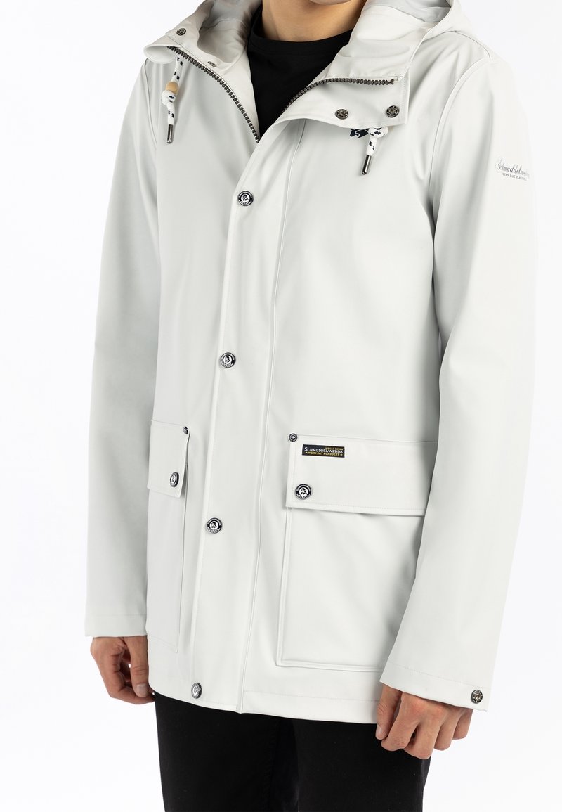 Homme portant une veste de pluie légère gris clair à capuche avec des boutons-pression à l'avant et deux grandes poches à rabat, chemise intérieure noire visible.