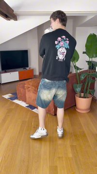 Svart hettegenser med grafikk av blomsterkrukke, slitte blå denimshorts og sølvfargede joggesko med svart tekst. Innendørs omgivelser med planter.