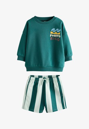 Kleines Kind mit geflochtenen Haaren steht und trägt einen grünen Pullover, grün-weiß gestreifte Shorts, grüne Socken und weiße Sneakers vor einem schlichten Hintergrund.