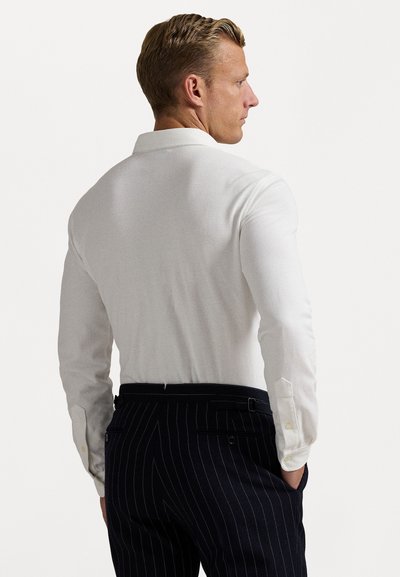 Cămașă albă cu mânecă lungă, dintr-un material texturat, cu un guler clasic și manșete cu nasturi, asortată cu pantaloni negri cu dungi.