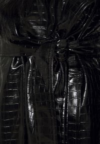 Czarny płaszcz z ekologicznej skóry o wzorze skóry krokodyla, wyposażony w szeroki pasek i błyszczące wykończenie. Widoczna teksturowana powierzchnia i szczegółowe szwy.