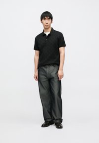 Polo nero con texture a motivi, abbinata a jeans grigio scuro. Il modello indossa scarpe nere. Design semplice e moderno.