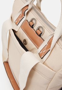 Bolsa de lona beige con detalles en cuero marrón, anillos de metal plateado, cierre con cremallera y bolsillo frontal con botón de presión magnético.