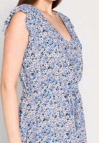 Blaues Blumenkleid mit Rüschensäumen, das kleine weiße, gelbe und orangefarbene Blumen auf einem hellblauen Hintergrund zeigt; verstellbarer Bindegürtel.