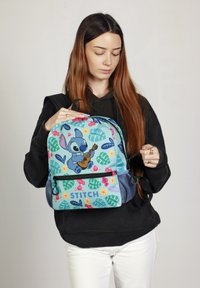 Mochila azul con un personaje de dibujos animados tocando la guitarra, con patrones florales y de hojas coloridos, detalles de malla y un bolsillo con cremallera en la parte delantera.