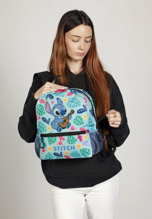 Mochila azul con un personaje de dibujos animados tocando la guitarra, con patrones florales y de hojas coloridos, detalles de malla y un bolsillo con cremallera en la parte delantera.