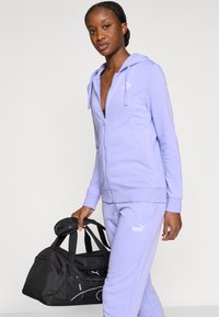 Λεβάντα ζακέτα με φερμουάρ και ταιριαστό παντελόνι φόρμας, και τα δύο από απαλό ύφασμα, με λευκές λεπτομέρειες λογότυπου Puma. Μαύρη σάκα με κοντά χερούλια.