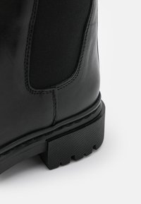 Musta nahkasaapas nilkkapituudella, jossa on matta pinta, joustavat sivupaneelit ja karkeapintainen kumipohja, jossa on selkeä kuviointi.